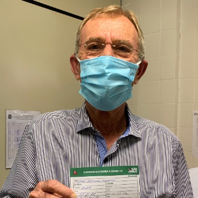 Foto de José Antonio Volpiani. Ele está de óculos e máscara, com semblante feliz, e segura o comprovante da vacinação contra a Covid-19